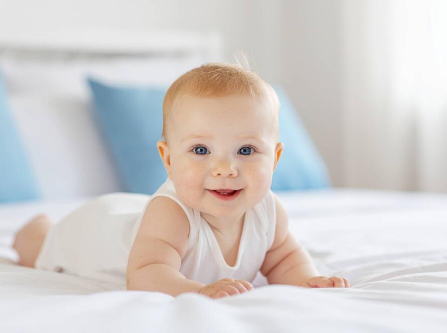 Services 6 Μωρό ξαπλωμένο σε λευκό κρεβάτι με χαμόγελο, σε φωτεινό υπνοδωμάτιο.