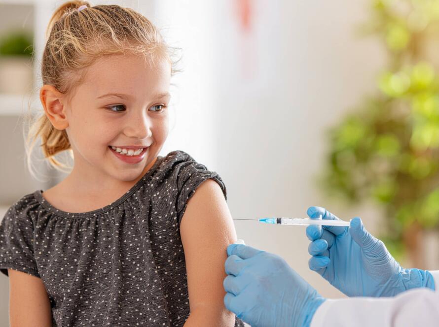 Services 2 Κοριτσάκι χαμογελά καθώς δέχεται εμβόλιο στο χέρι από γιατρό με γάντια, σε ιατρικό περιβάλλον.