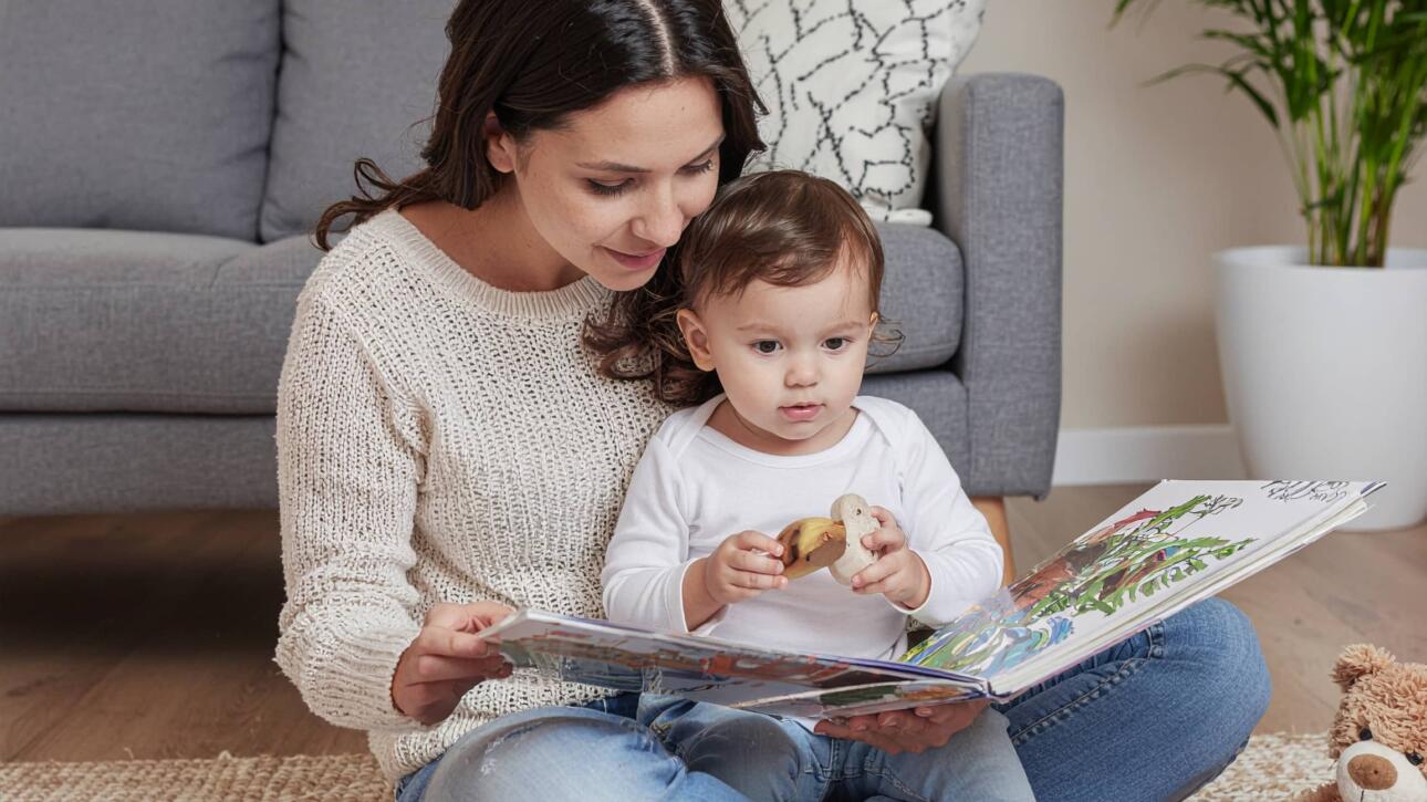 Η σημασία της ανάγνωσης από την πρώτη βρεφική ηλικία 3 Μαμά διαβάζει βιβλίο σε μικρό παιδί καθισμένες στο πάτωμα του σαλονιού, με λούτρινα παιχνίδια δίπλα τους