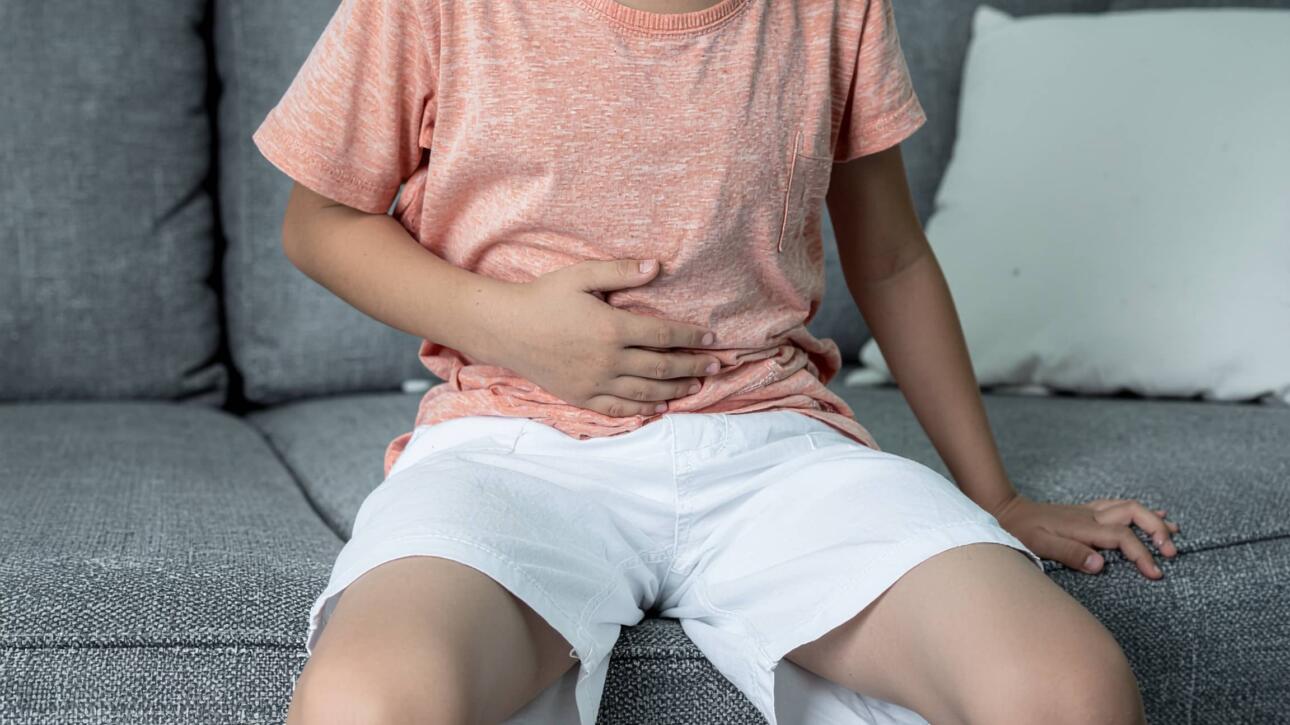 Εντεροϊοί στα παιδιά: πώς να αναγνωρίσετε τη λοίμωξη & πότε να ανησυχήσετε; 2 Αγόρι με πορτοκαλί μπλούζα και λευκό σορτς κάθεται σε καναπέ και κρατά την κοιλιά του, δείχνοντας ενόχληση ή πόνο.