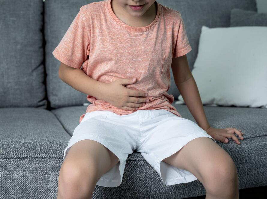 Αγόρι με πορτοκαλί μπλούζα και λευκό σορτς κάθεται σε καναπέ και κρατά την κοιλιά του, δείχνοντας ενόχληση ή πόνο.