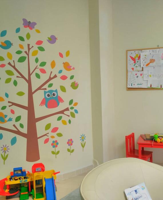 Τοίχος παιδικού χώρου διακοσμημένος με αυτοκόλλητο δέντρο και πουλάκια, παιδικό τραπεζάκι με κόκκινες καρέκλες και παιχνίδια, και πίνακας με ζωγραφιές παιδιών