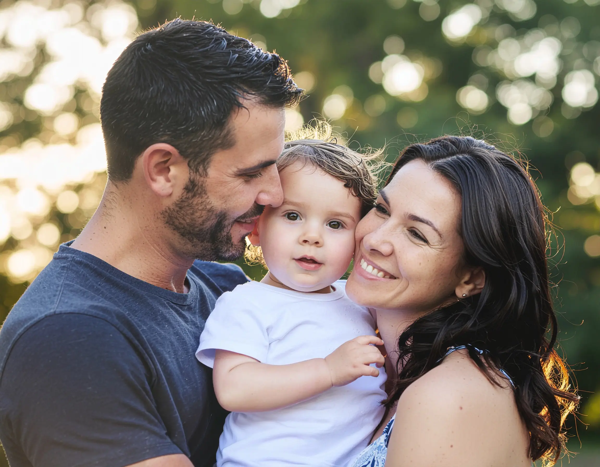 Νεαροί γονείς αγκαλιάζουν το παιδί τους σε εξωτερικό χώρο με ηλιόλουστο φόντο, εκφράζοντας αγάπη και οικογενειακή θαλπωρή