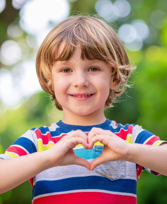 Αγόρι με πολύχρωμη μπλούζα κάνει σχήμα καρδιάς με τα χέρια του και χαμογελά σε εξωτερικό χώρο με πράσινο φόντο